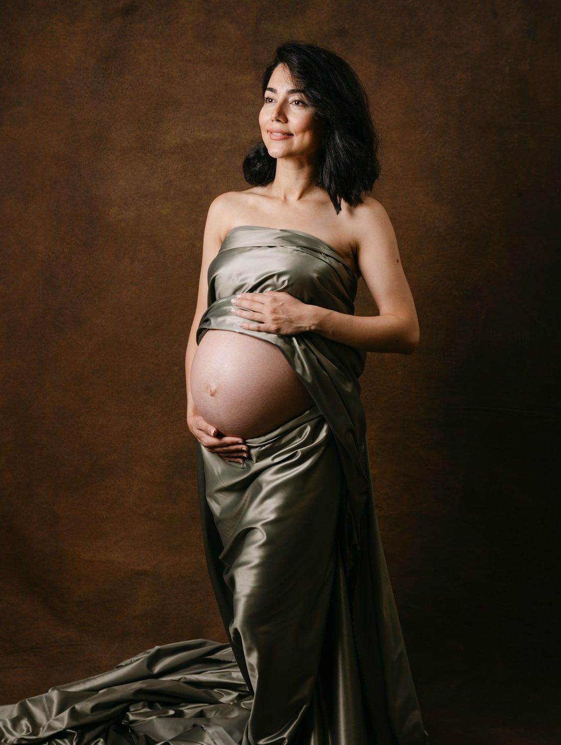 Pregnant woman draped in olive satin fabric poses elegantly against a brown studio backdrop for maternity portrait.
