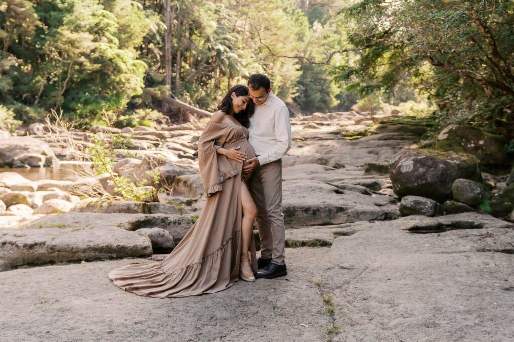 Couple posing on rocky terrain in a lush forest, woman wearing an elegant flowing tan maternity gown.