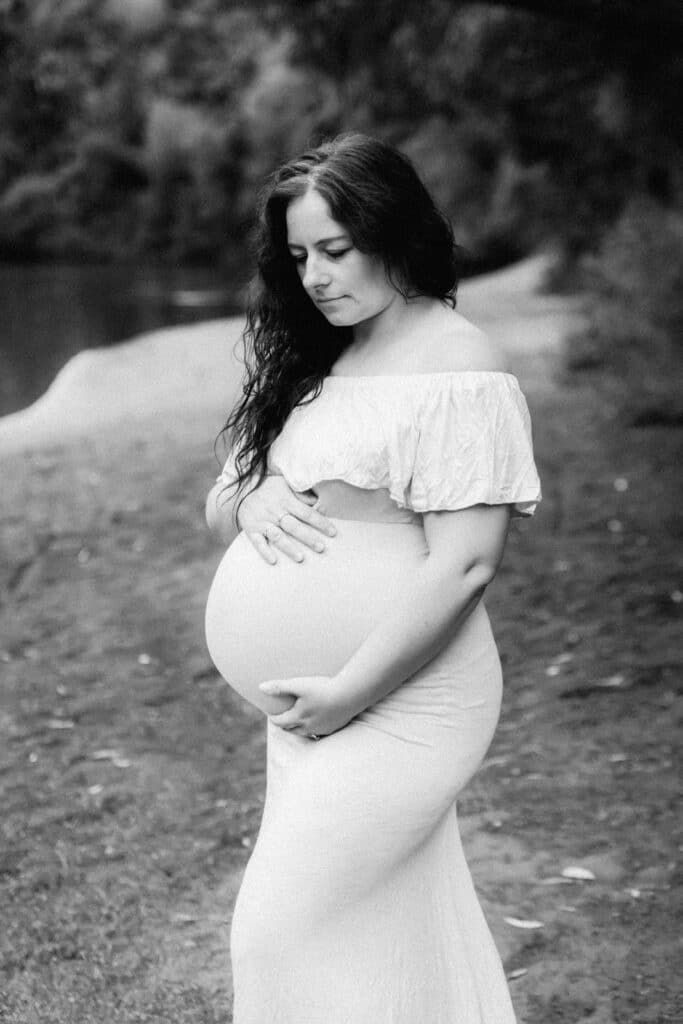 Black and white maternity photo of pregnant woman in off-shoulder dress holding her baby bump outdoors.