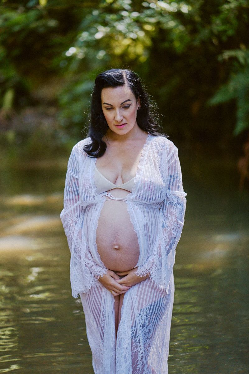 Pregnant woman in white lace robe standing in water, cradling her baby bump, surrounded by lush green trees.