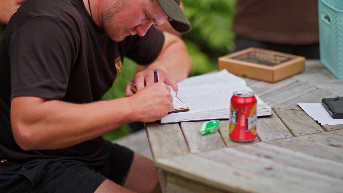 A UPS delivery worker in brown uniform writes on clipboard at outdoor wooden table with Coca-Cola can nearby.