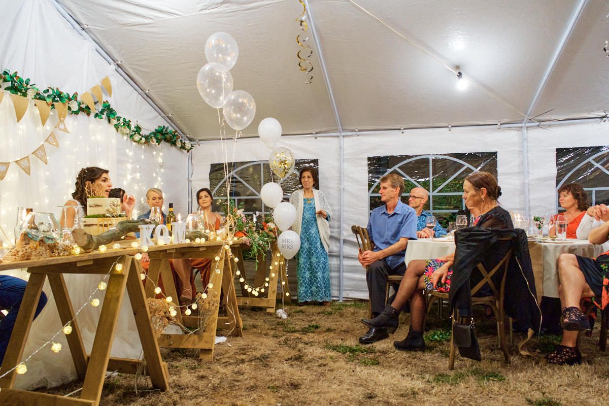 Elegant outdoor wedding reception under a white tent with guests seated at decorated tables with string lights and balloons.