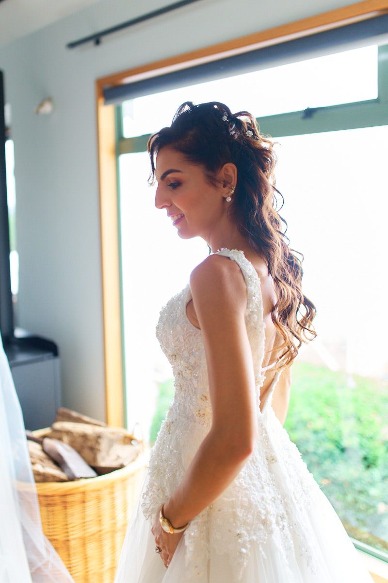 Bride in lace wedding dress standing by window, smiling softly with elegant half-up hairstyle and gold bracelet.