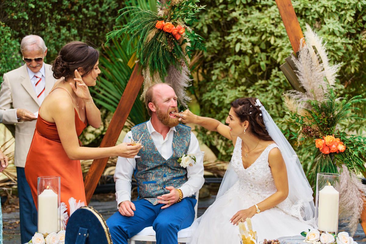 Bride feeds groom cake during outdoor wedding ceremony with bridesmaid and officiant in lush garden setting.