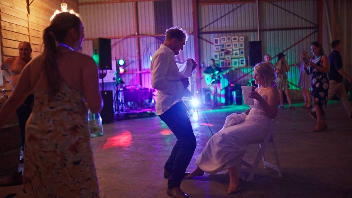 Bride and groom dancing joyfully at a rustic barn wedding reception with colorful lights and live band.