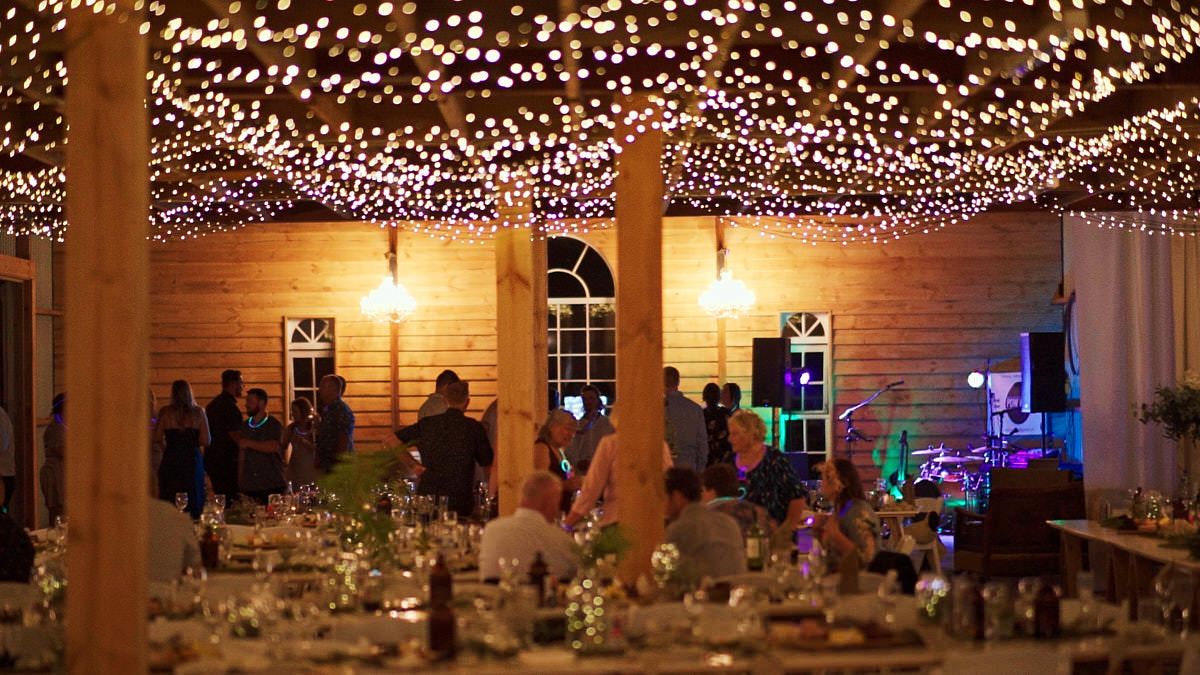 Elegant outdoor wedding reception with string lights canopy, decorated tables, and guests mingling at a warmly lit venue.