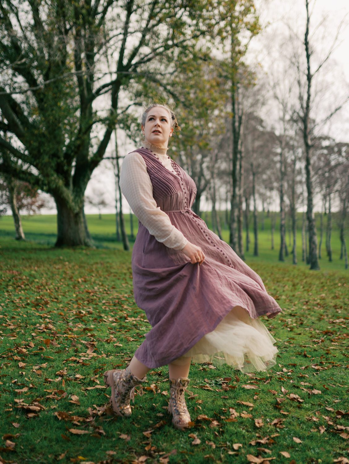 Woman in purple dress and lace boots twirling outdoors among autumn trees on green grass.
