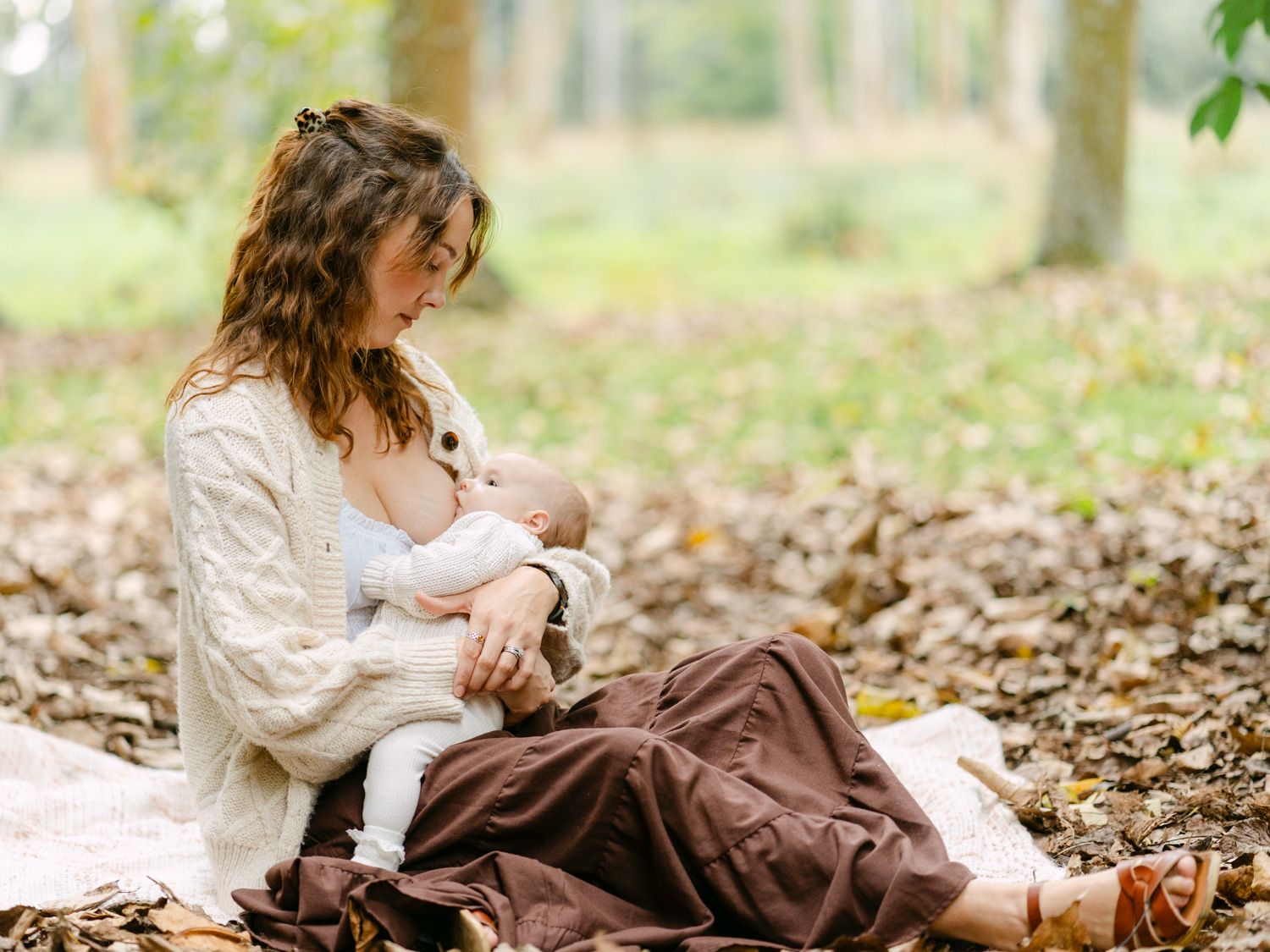 Mother breastfeeding newborn baby outdoors in autumn forest, sitting on blanket among fallen leaves.
