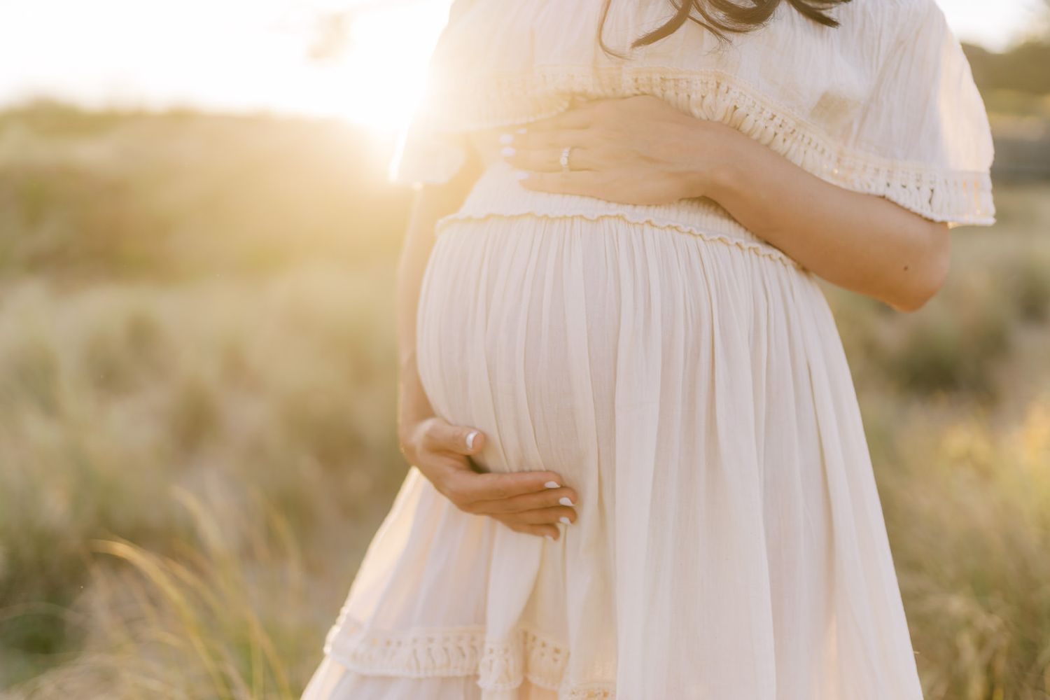 Pregnant woman in white dress cradling baby bump in golden sunset field.