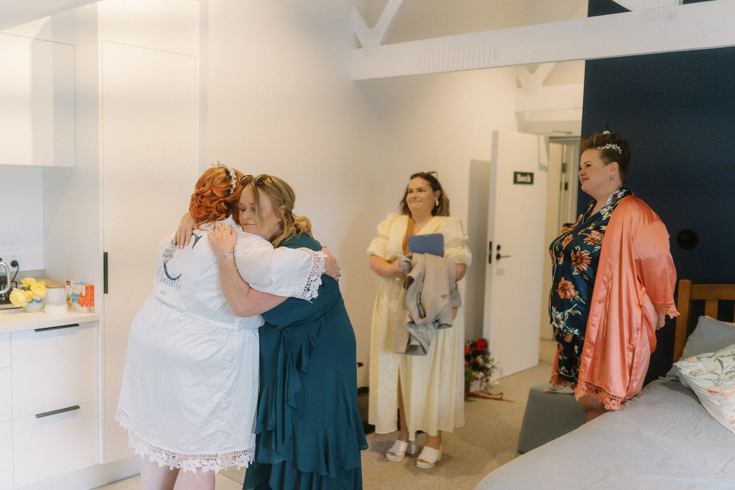 Women in colorful dresses hugging and celebrating together in a bright modern room during a wedding preparation.