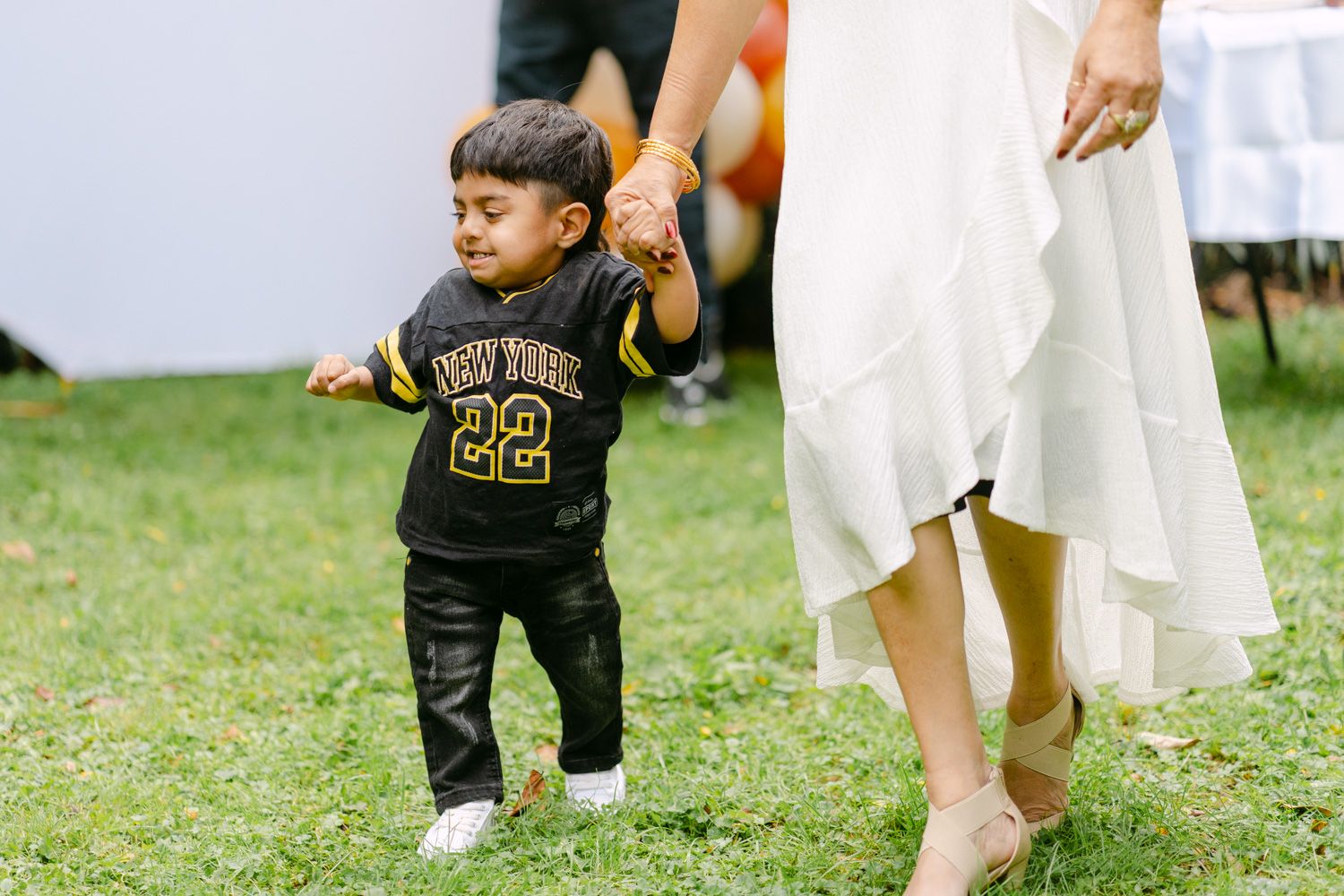 Toddler in New York 22 jersey walks on grass holding adult's hand at outdoor event.