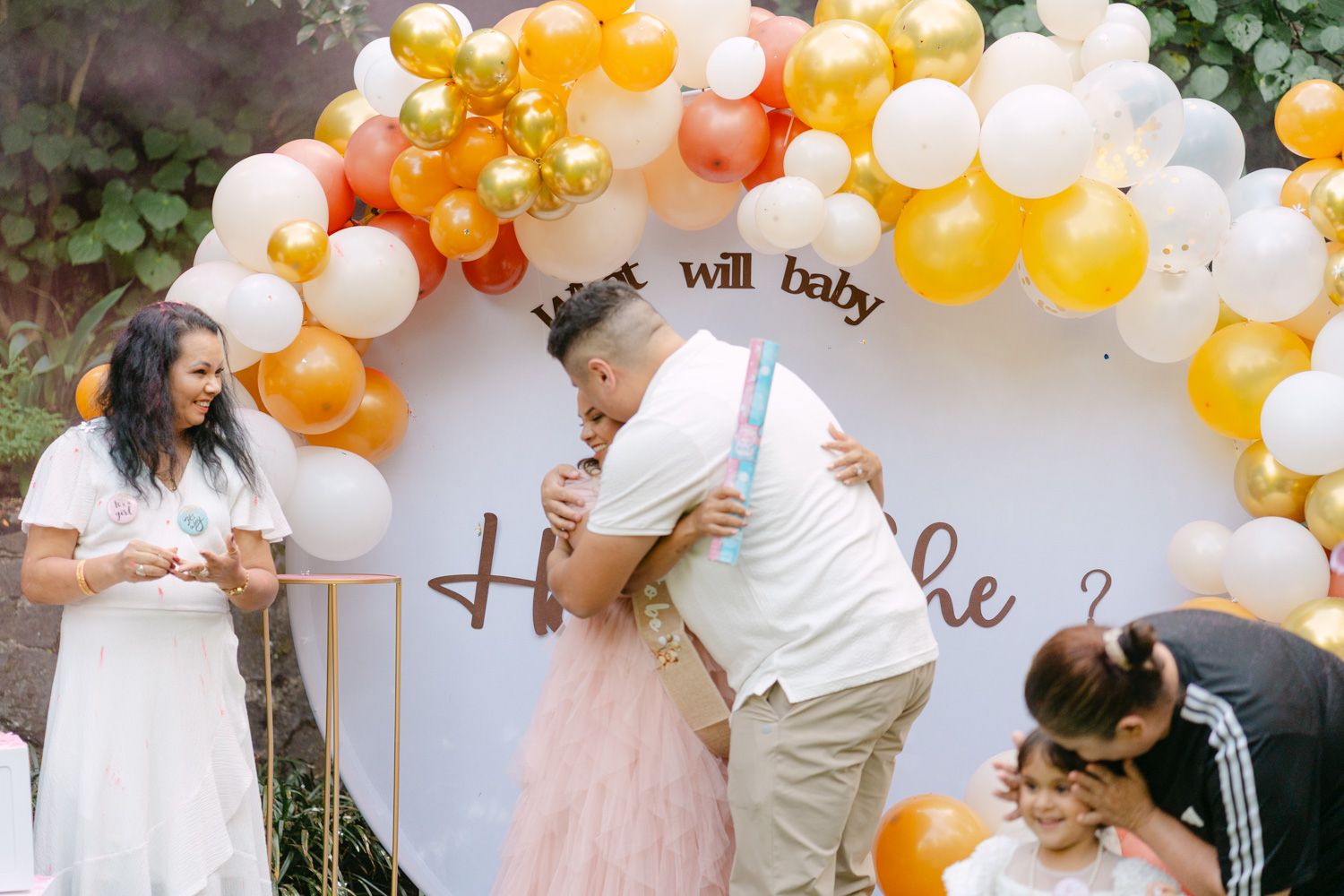 Couple embracing at a gender reveal party under a colorful balloon arch with the text 'What will baby be?'.