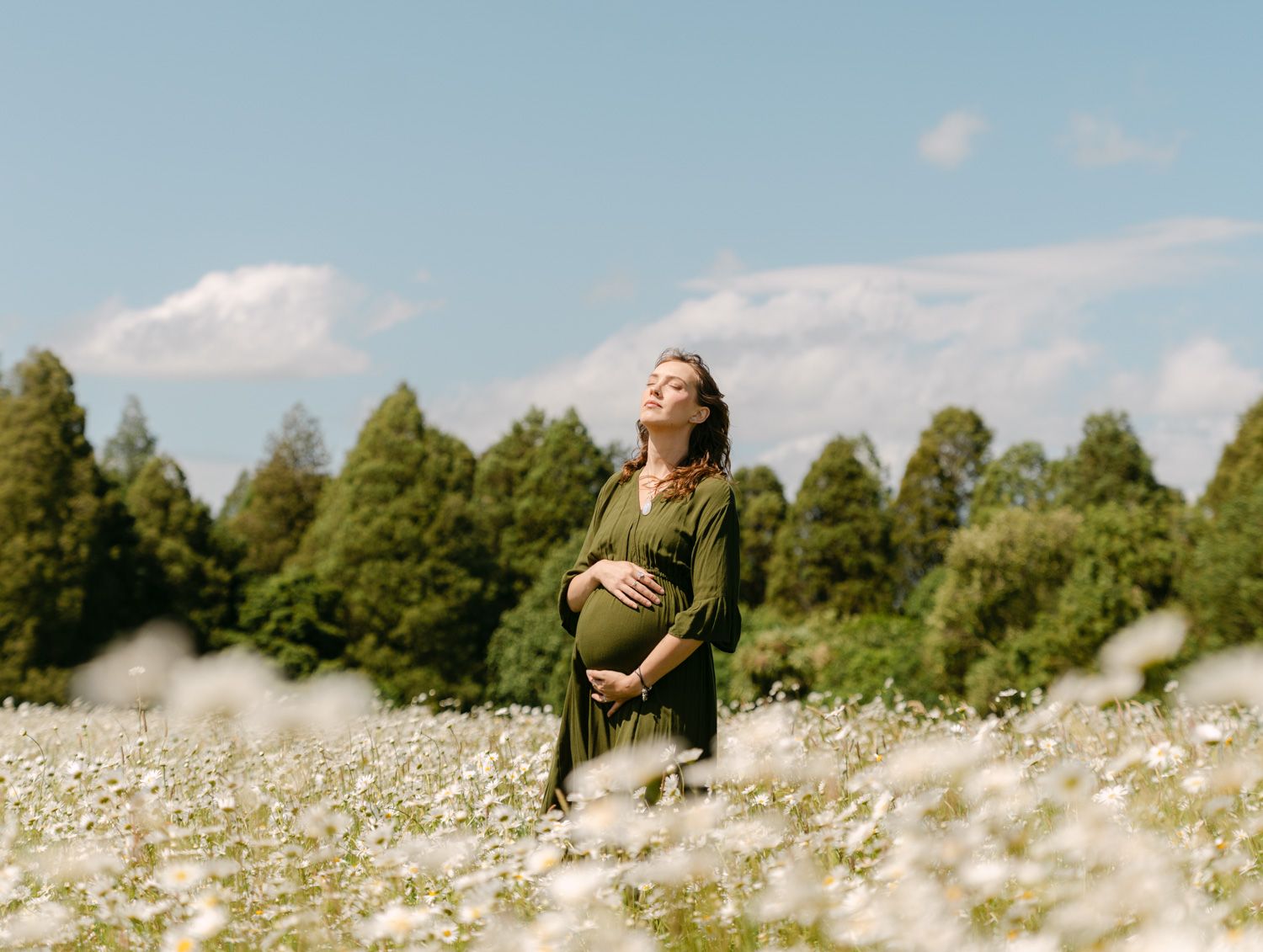 Pregnant woman in green dress standing in white wildflower field, hands on belly, trees and blue sky behind her.