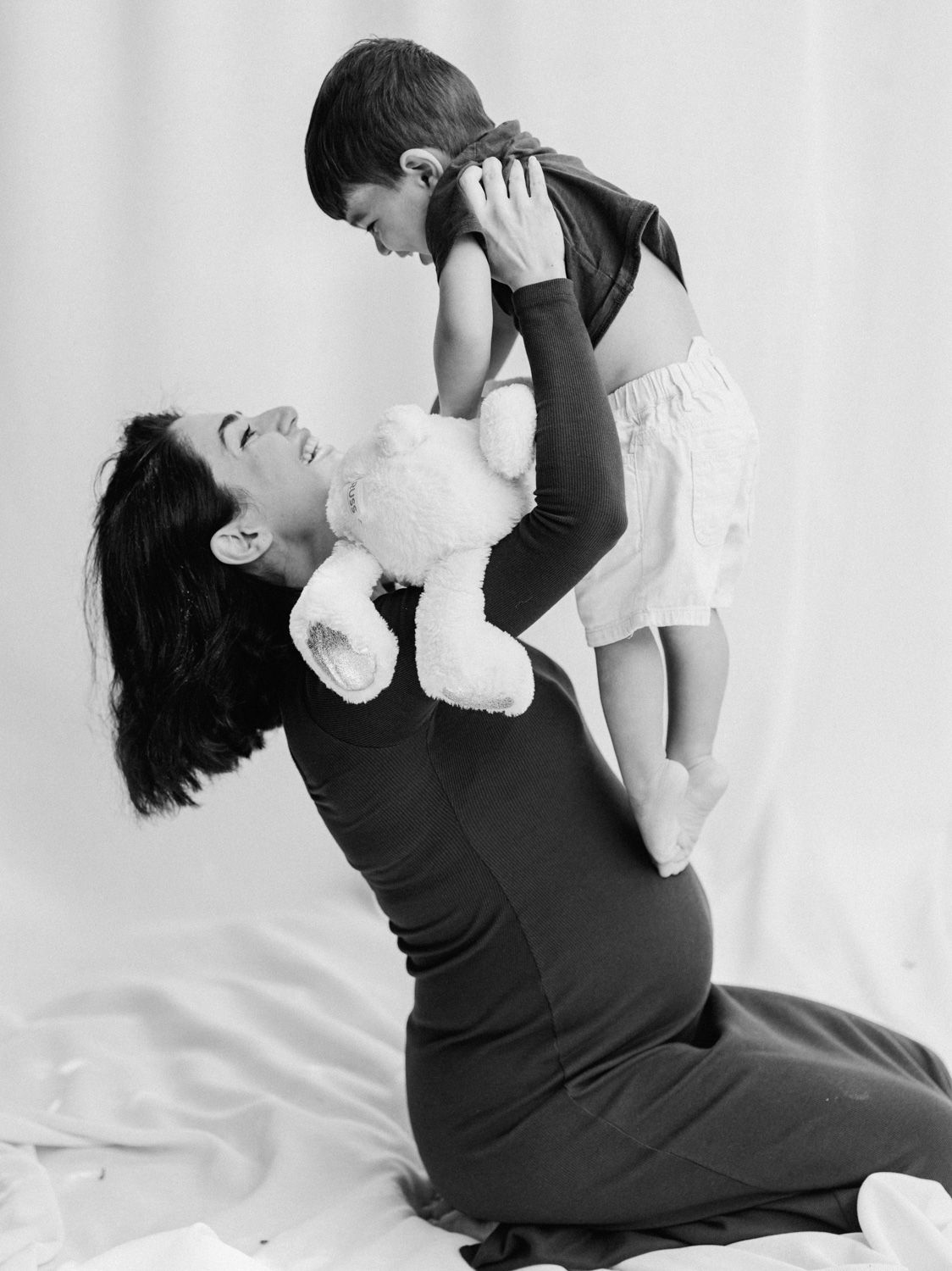 Black and white photo of a mother kneeling, lifting her toddler up while holding a teddy bear, sharing a joyful moment.