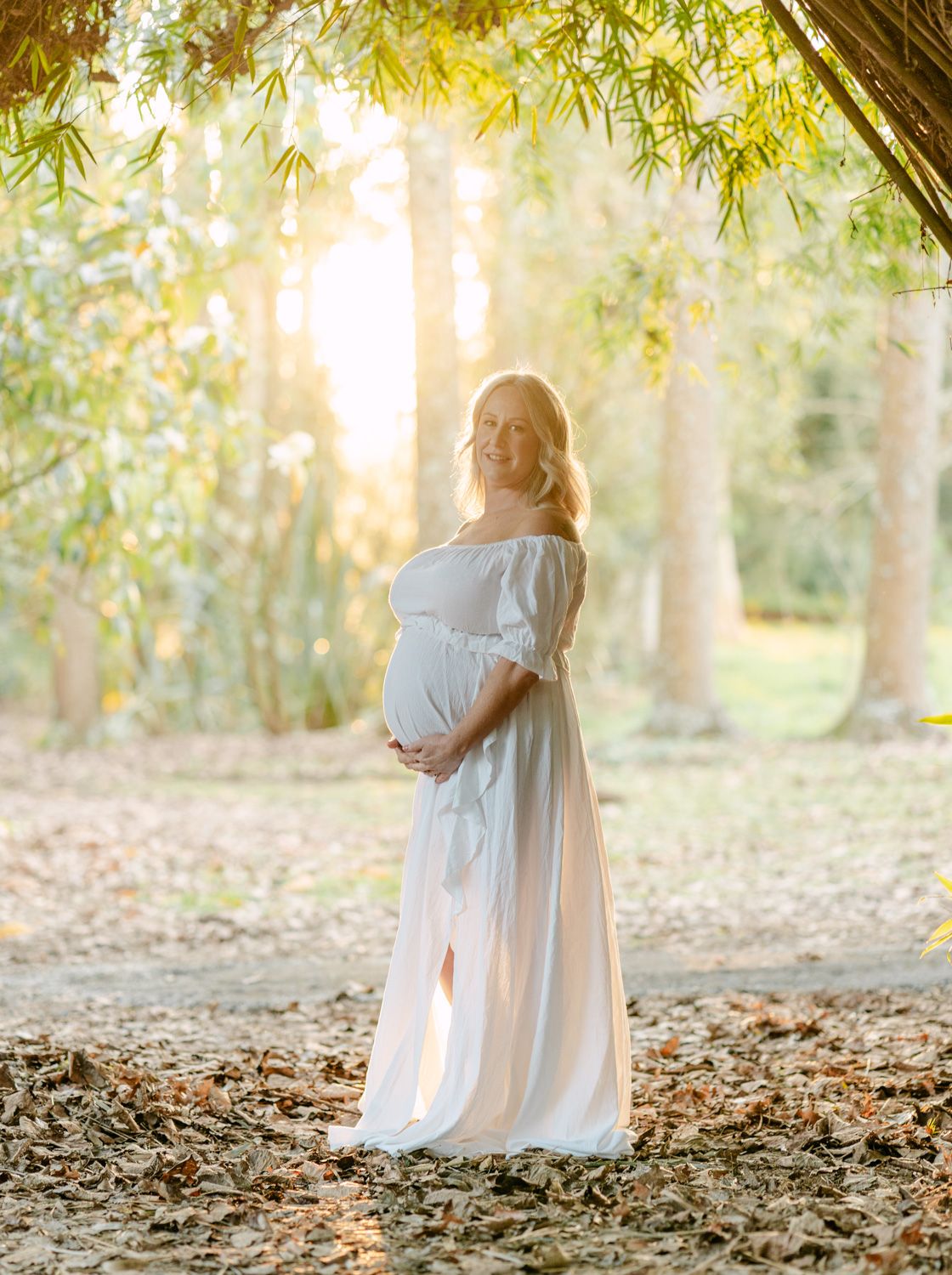 Pregnant woman in white off-shoulder gown standing among trees in golden sunlight, cradling her baby bump.