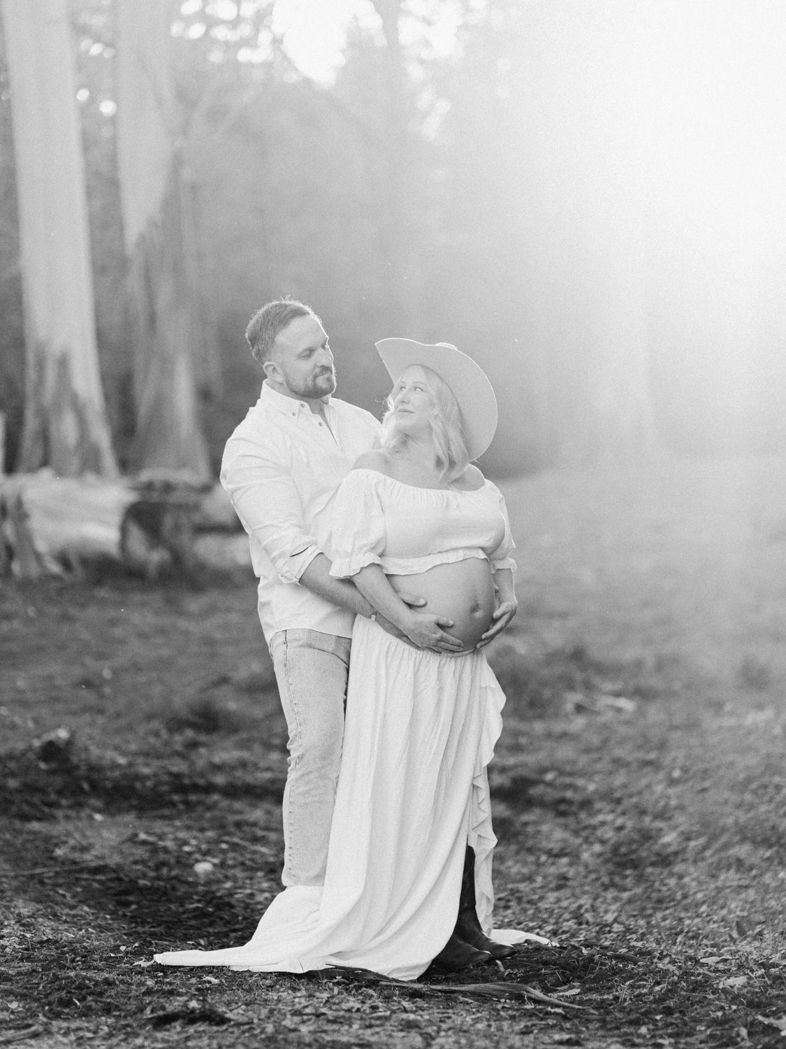 Black and white maternity photo of couple embracing outdoors, pregnant woman in flowing dress and hat, sun flare background.