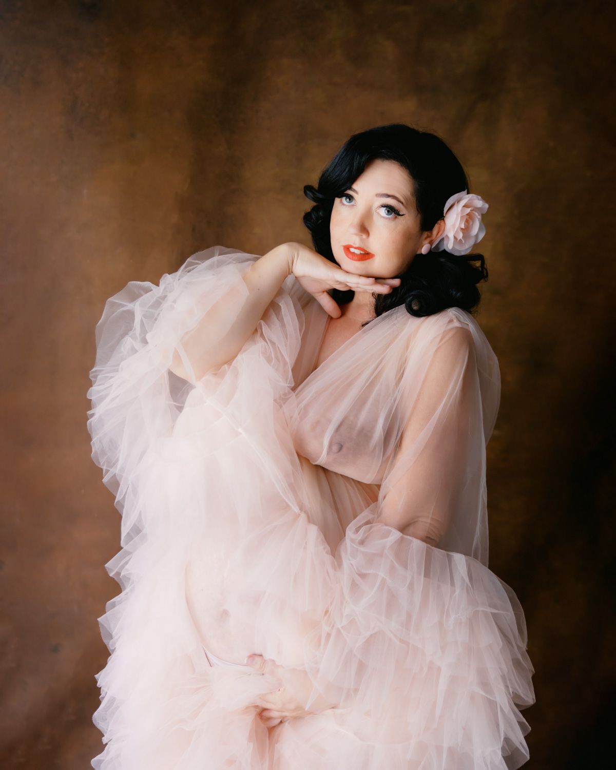 Woman in pink tulle gown with floral hair accessory poses gracefully against brown backdrop.