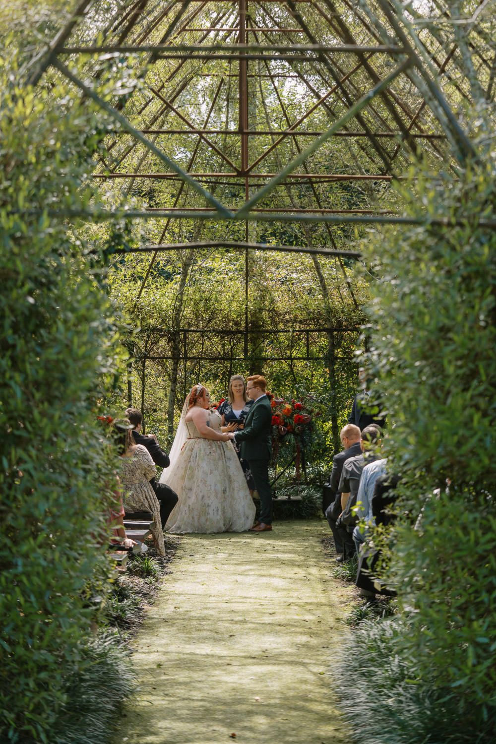 Couple exchanges vows in a lush green garden gazebo wedding ceremony with guests seated along a leafy aisle.
