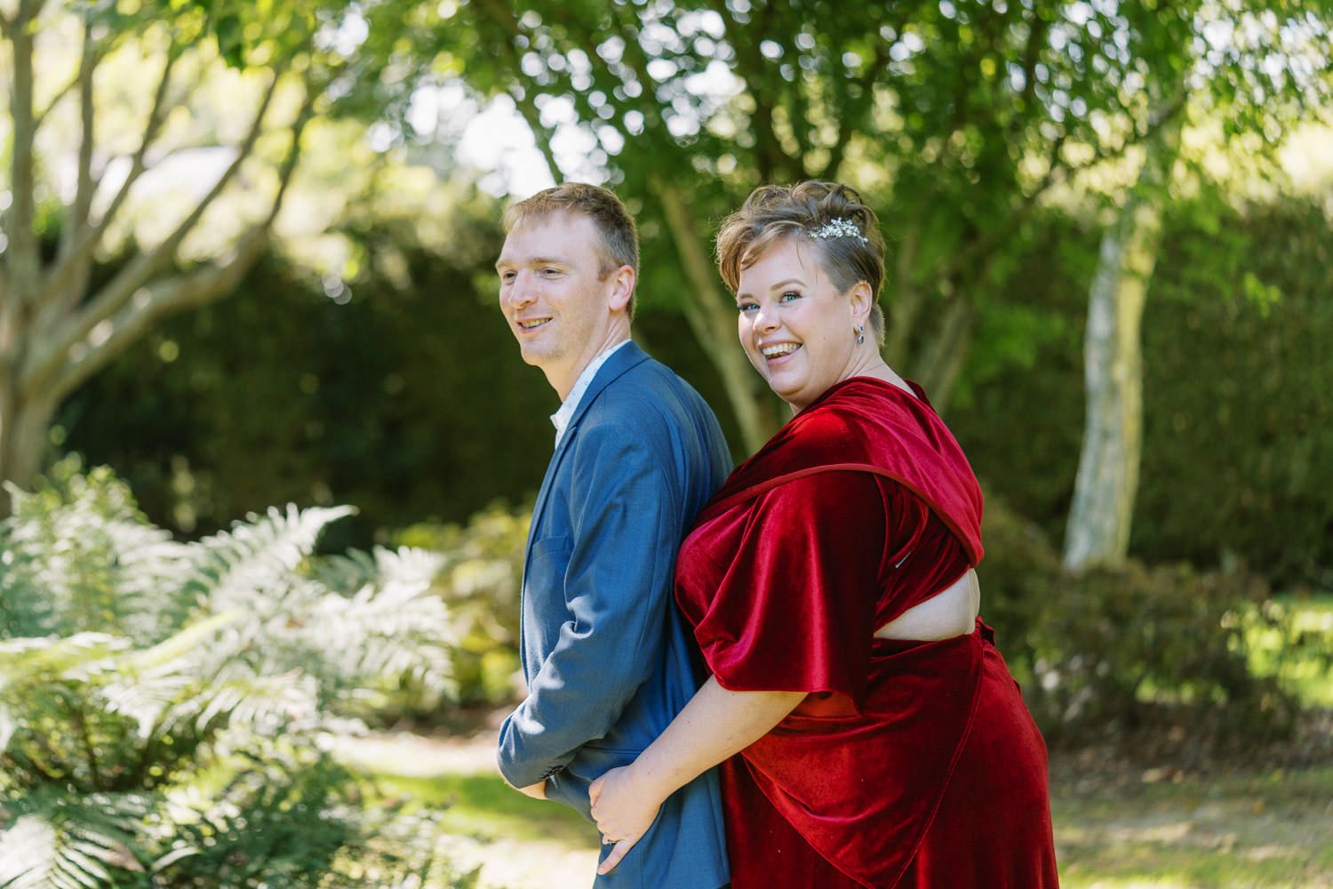 Couple posing back-to-back outdoors, man in blue suit and woman in red velvet gown, surrounded by lush green trees.