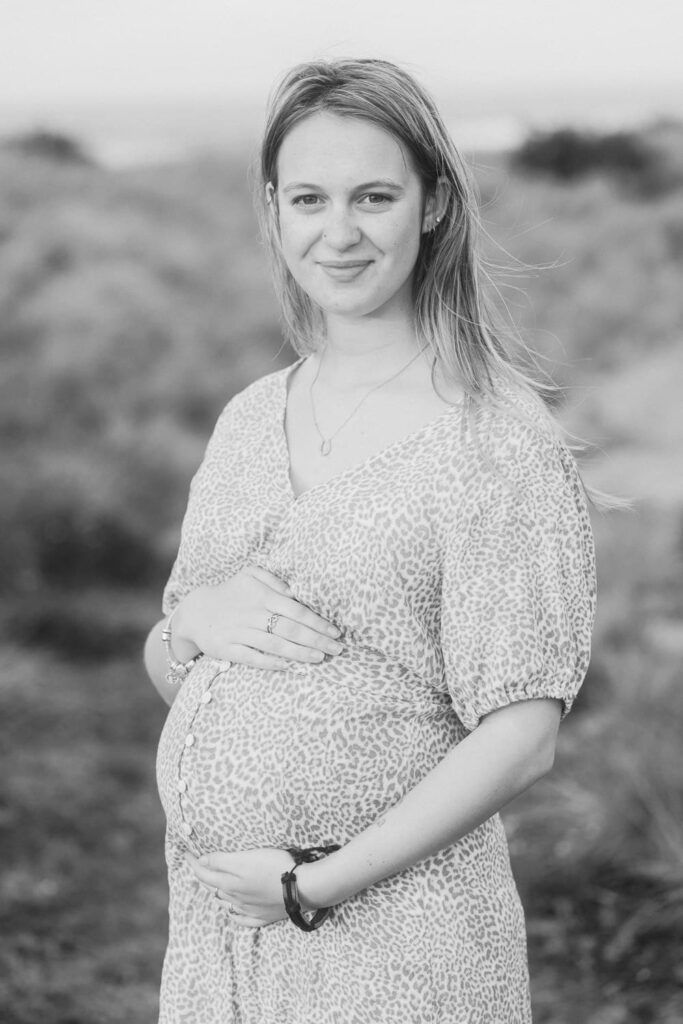 Black and white portrait of a smiling pregnant woman in a floral dress, cradling her baby bump outdoors.