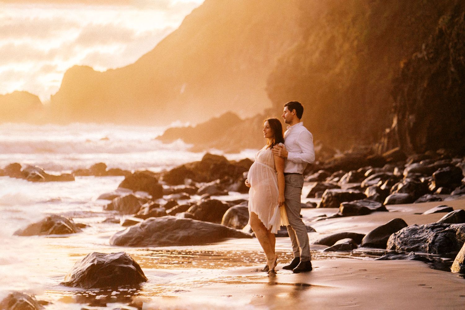 Couple embracing on a rocky beach at golden sunset with dramatic coastal cliffs and glowing ocean waves.