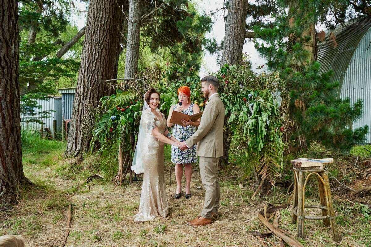 Outdoor woodland wedding ceremony with bride in ivory gown, groom in tan suit, and officiant in floral dress among pine trees.