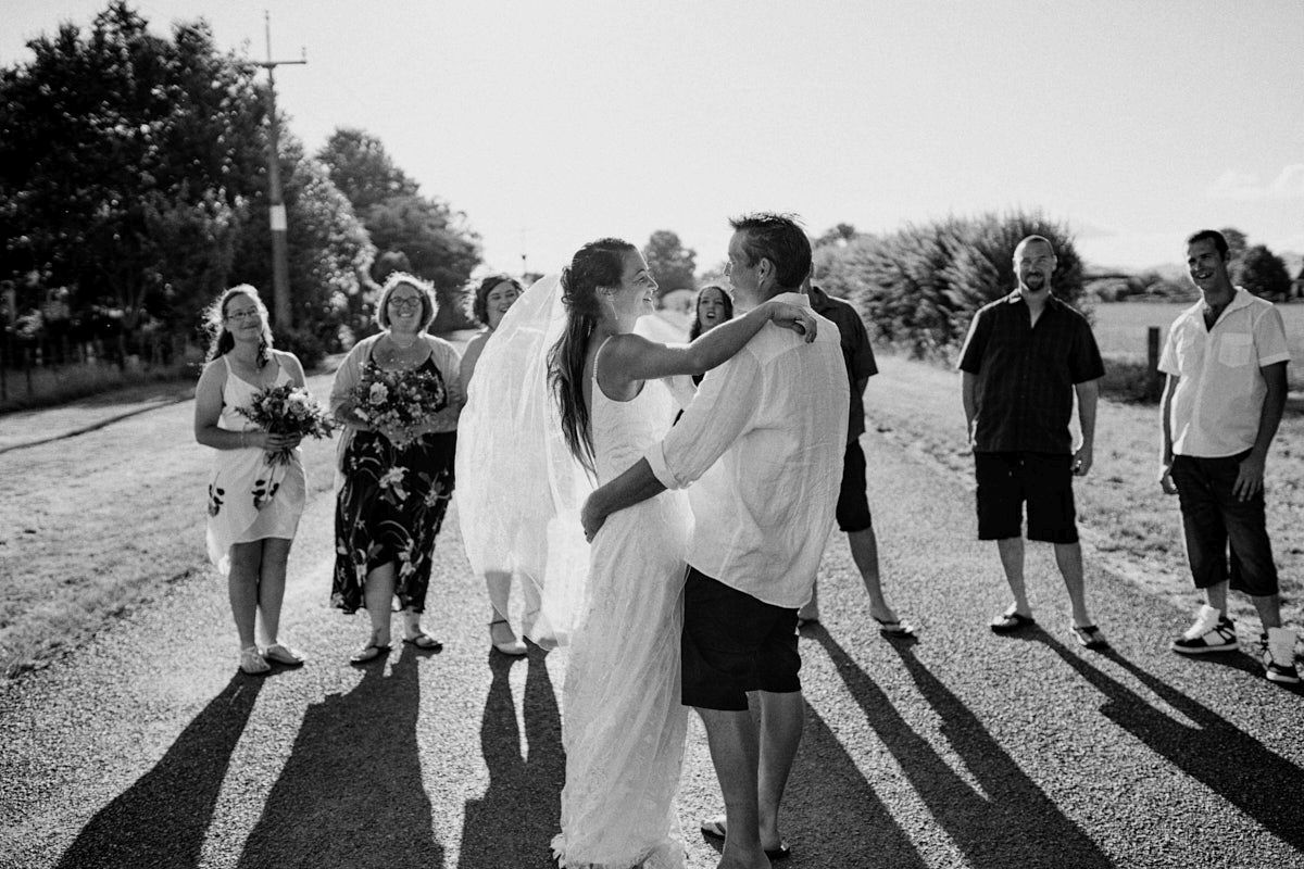 Black and white photo of a bride and groom sharing a kiss outdoors, surrounded by wedding guests on a sunny day.