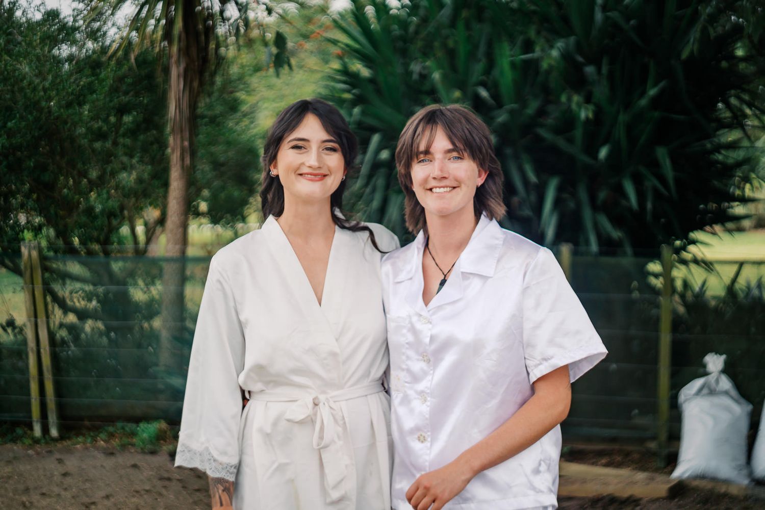 Two smiling women in white robes standing outdoors surrounded by lush green trees and plants.