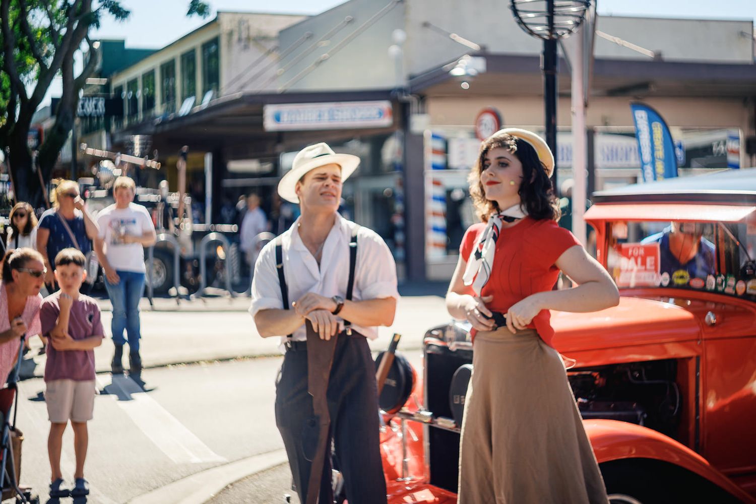 Two people in 1940s vintage attire pose near a classic red car on a sunny street during a retro festival.