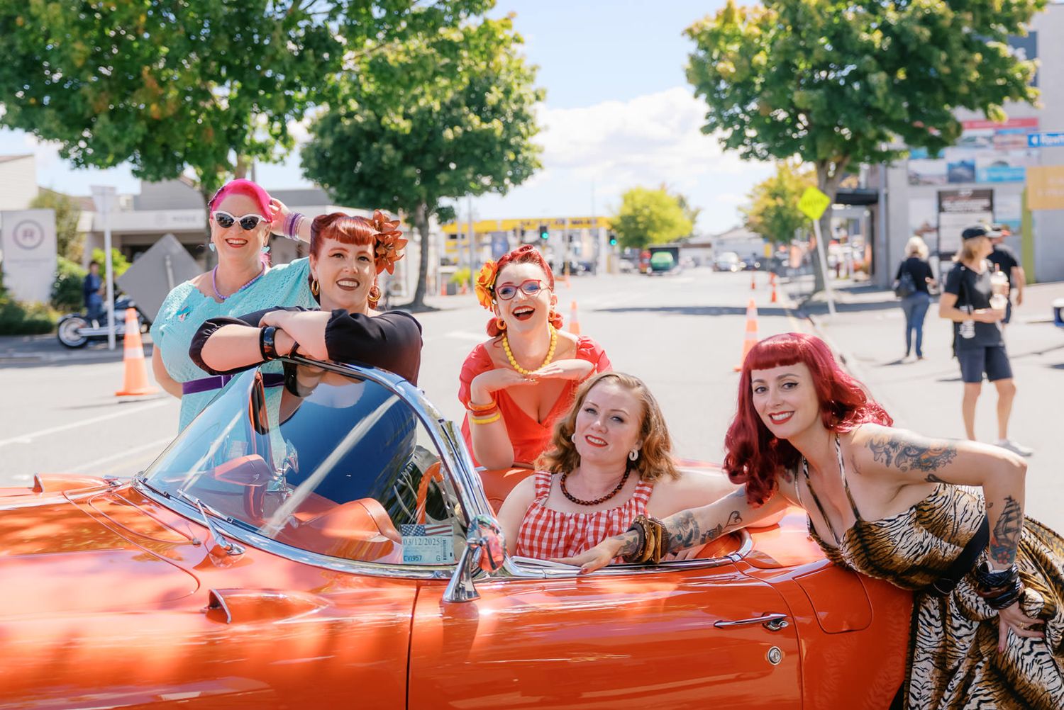 Five retro-styled women in vintage pin-up outfits posing in a classic orange convertible car on a sunny street.