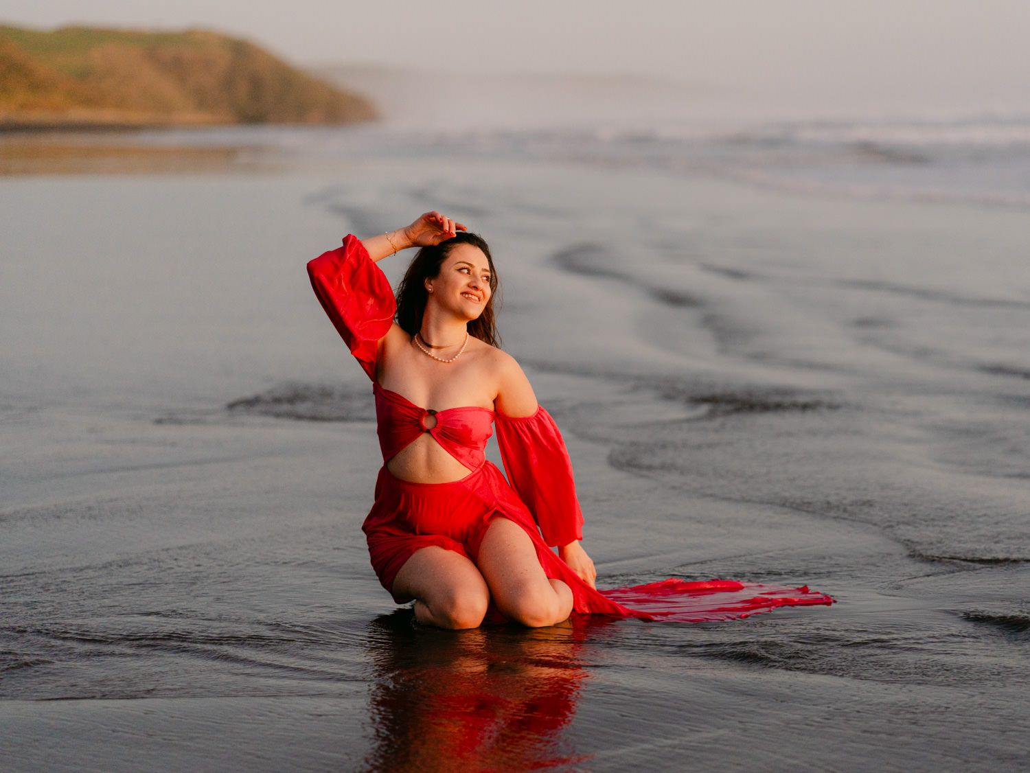 Woman in red bikini and sheer wrap posing on a wet beach at sunset with coastal cliffs in background.