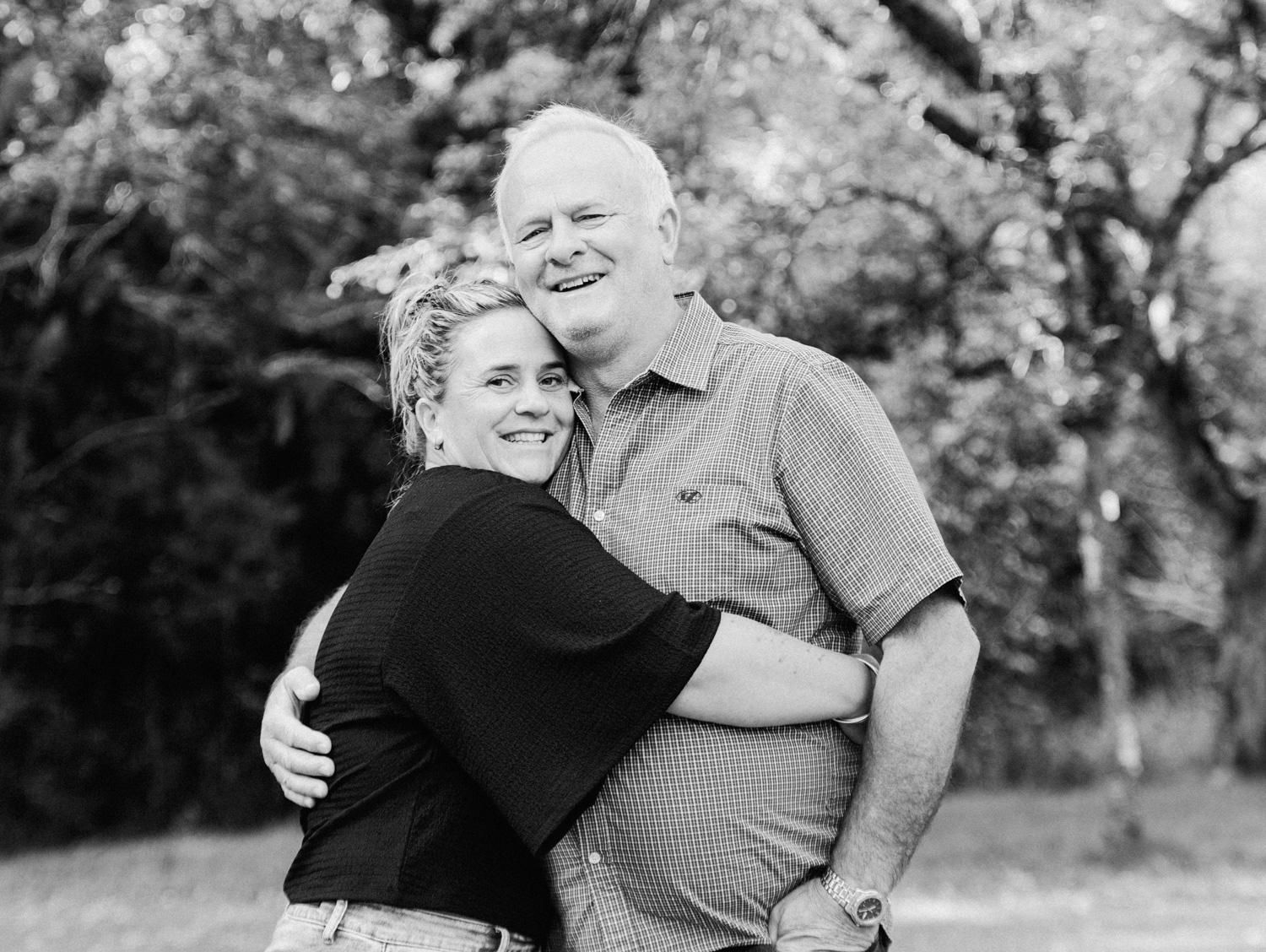 Black and white portrait of a smiling older man and young woman hugging outdoors among trees.