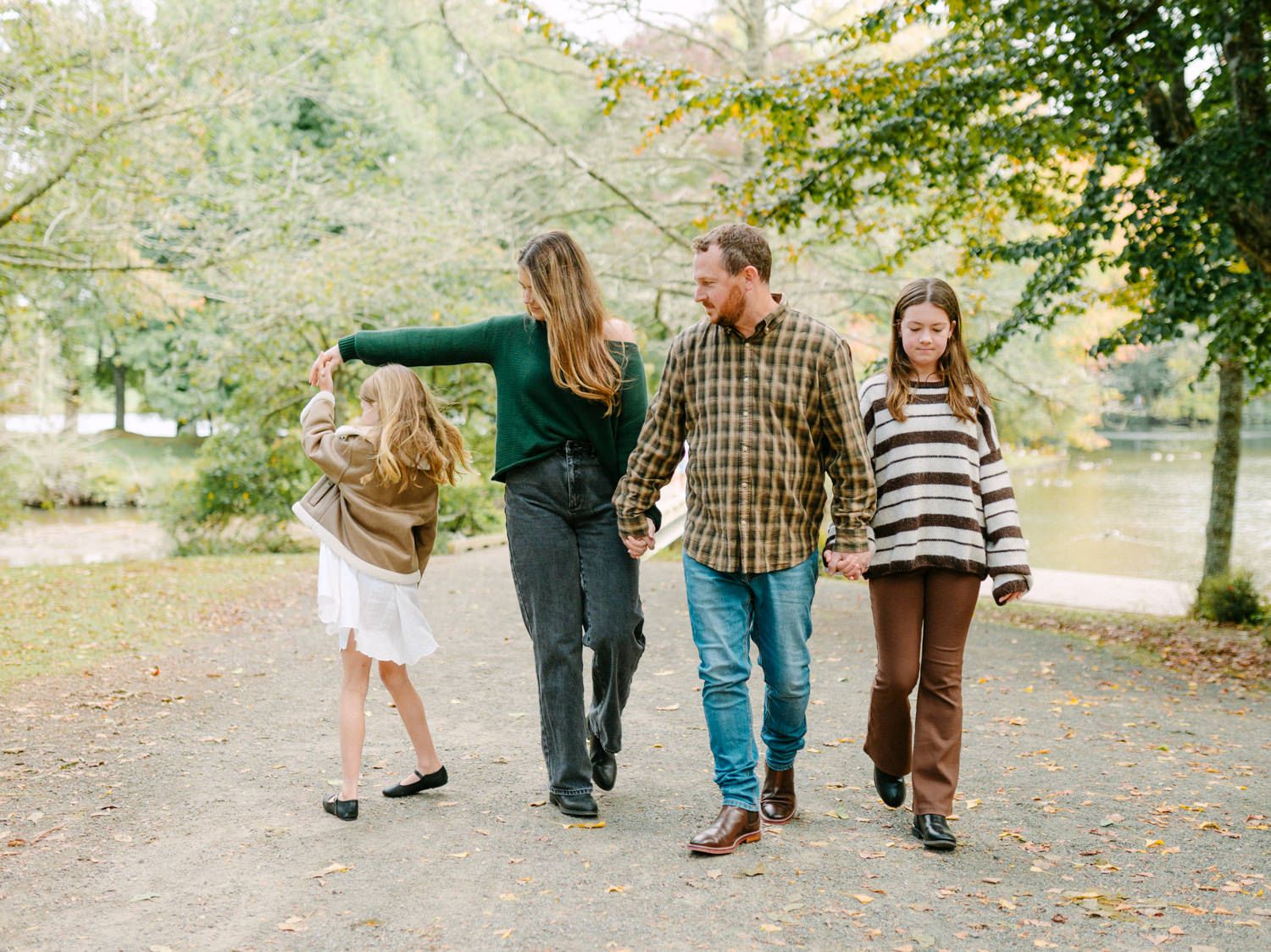 Family of four walking hand-in-hand on a scenic park path surrounded by autumn foliage near a pond.