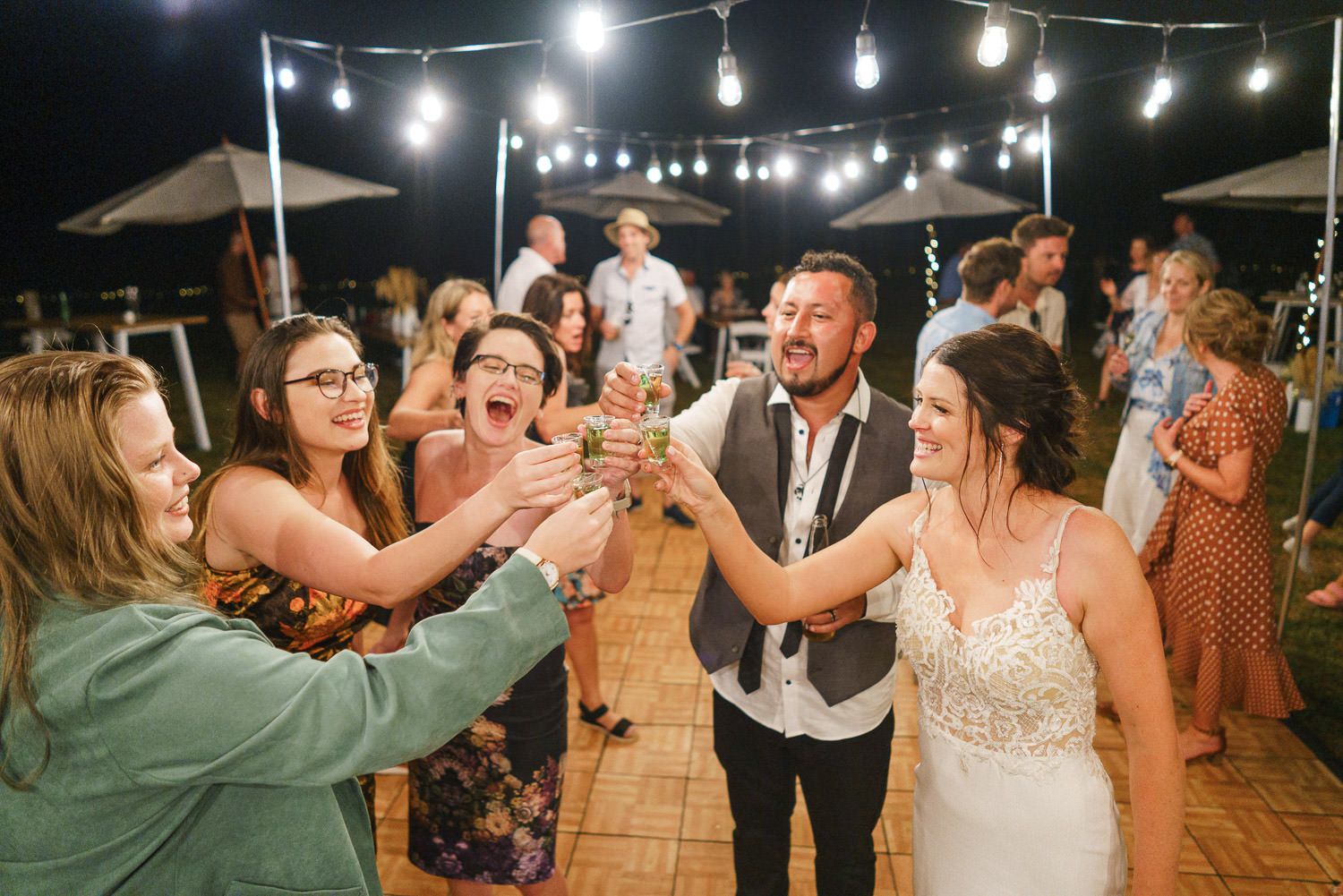Wedding party toasting drinks outdoors at night under string lights.