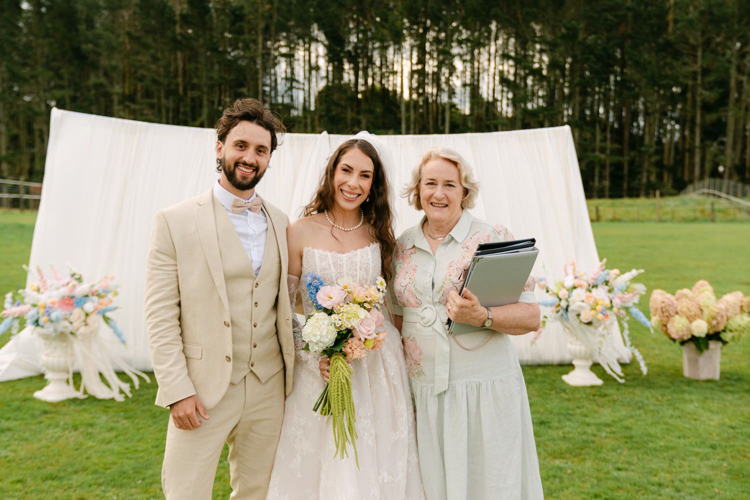 Bride and groom pose with wedding officiant outdoors, white drape backdrop, colorful floral arrangements on green lawn.