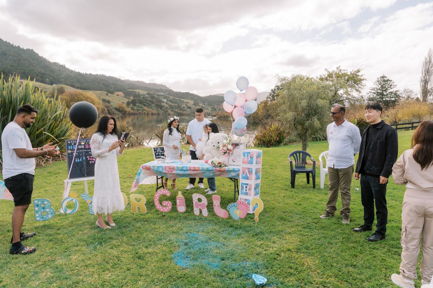 A gender reveal party on a green lawn with guests gathered around pink decorations and balloons, set against a scenic mountain backdrop.
