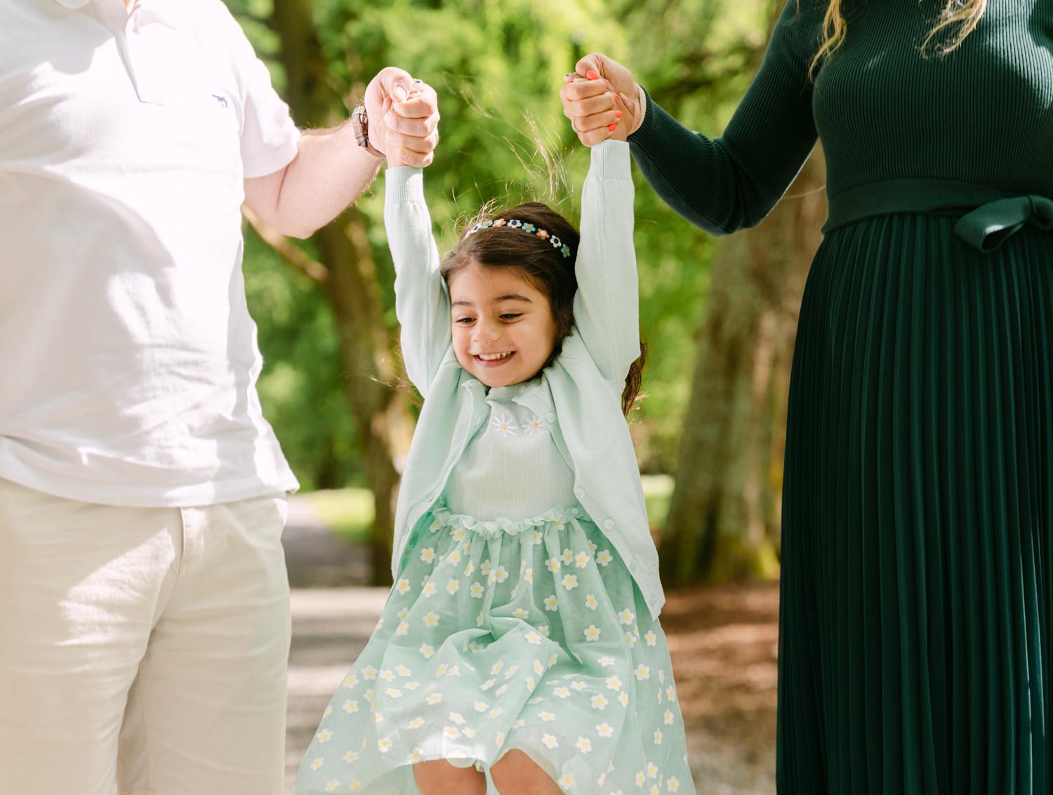 Happy little girl in mint dress swings between parents holding her hands in a sunny park.