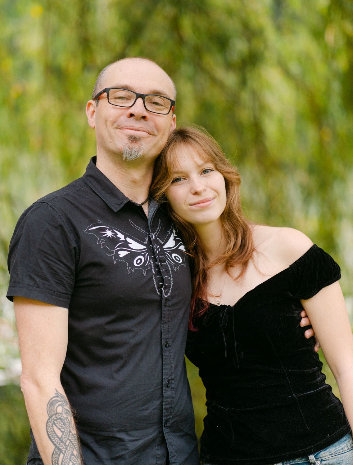 A bald man with glasses and tattoos poses with a young woman in a black top, smiling outdoors near green trees.