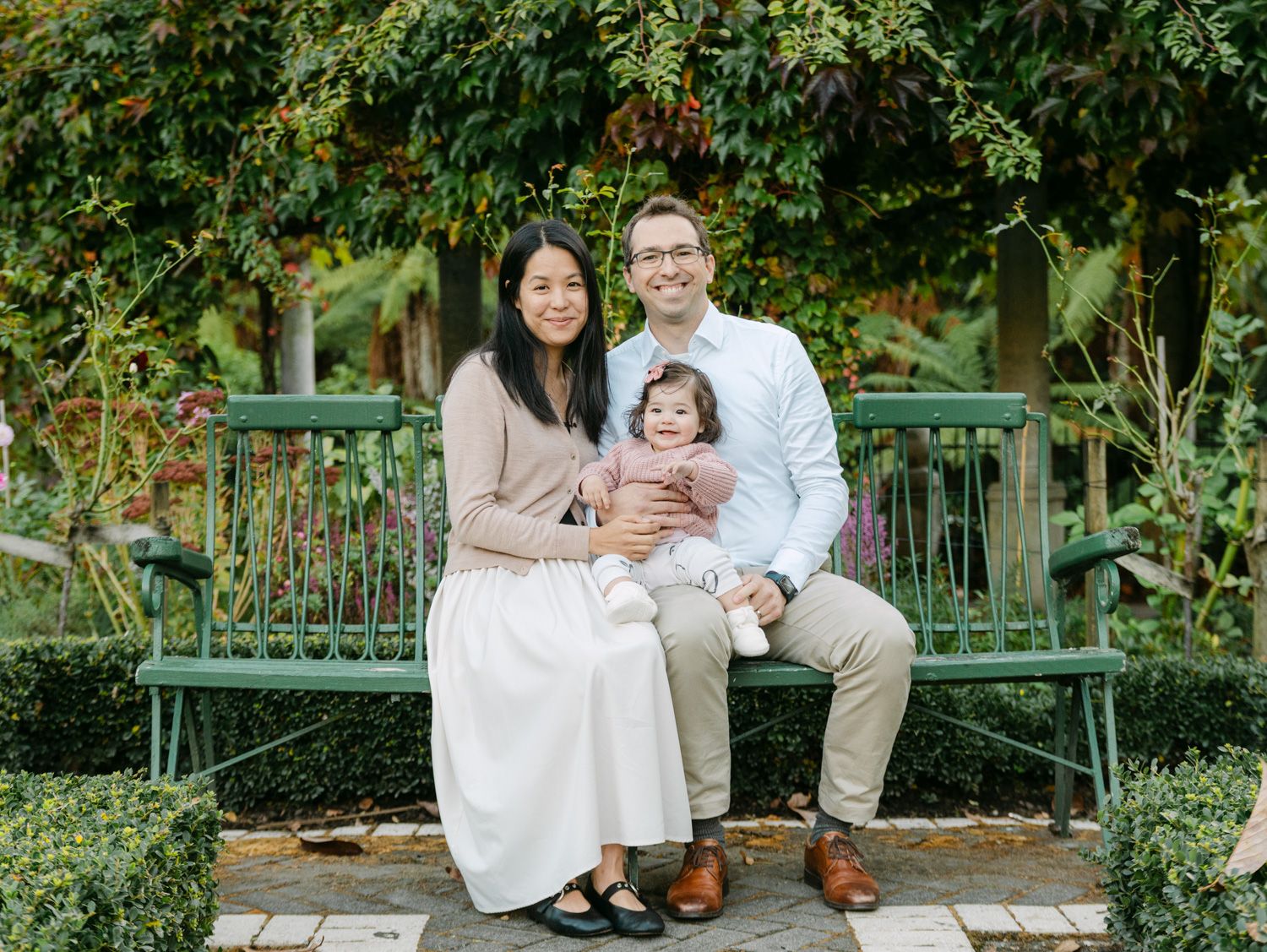 A happy family of three poses on a green garden bench surrounded by lush greenery.