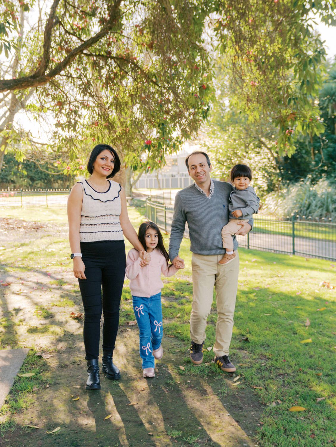A happy family of four walks through a sunny park, parents holding children's hands under a leafy tree.