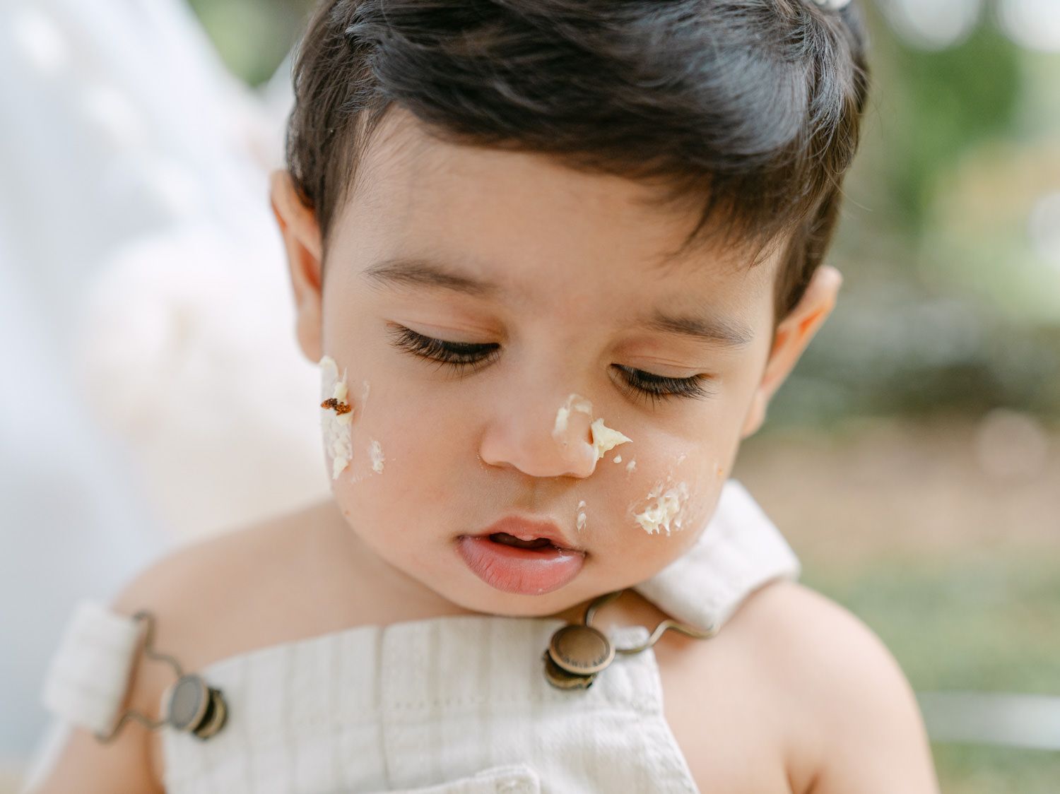 Adorable baby with cake on face wearing white overalls during first birthday smash cake photo session.