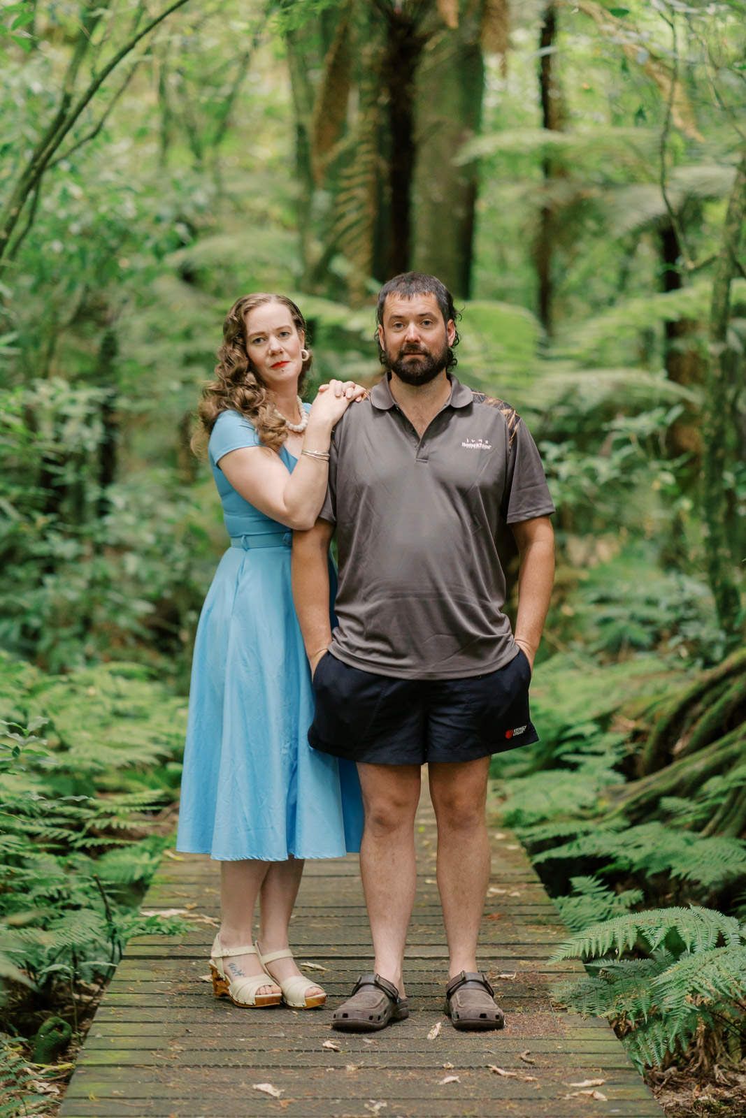 Couple standing on wooden boardwalk in lush green forest, woman in blue dress, man in grey polo shirt.