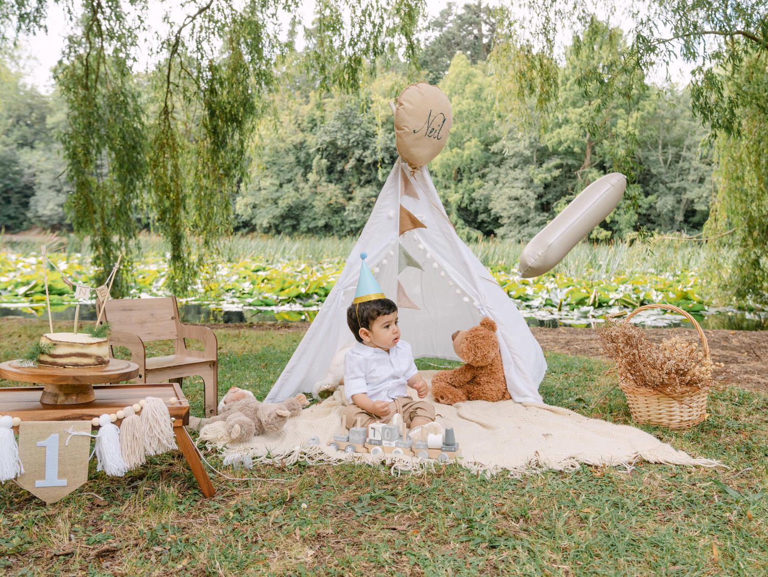 Toddler boy sitting beside a large teddy bear in front of a white teepee tent in a lush garden setting.