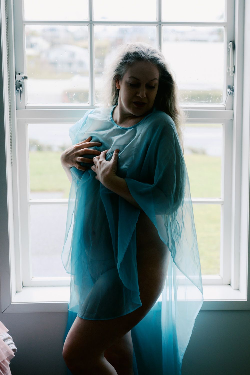 Woman in sheer teal wrap standing by a bright window in a soft, artistic boudoir-style portrait.
