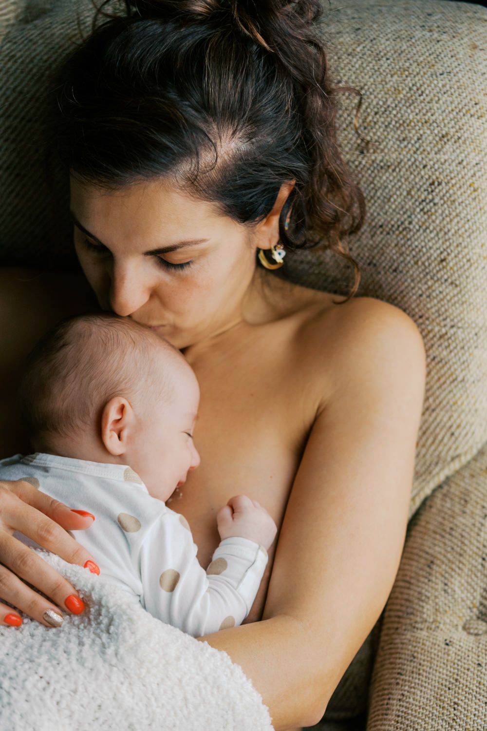 A mother tenderly kisses her newborn baby while cradling them against her chest on a couch.