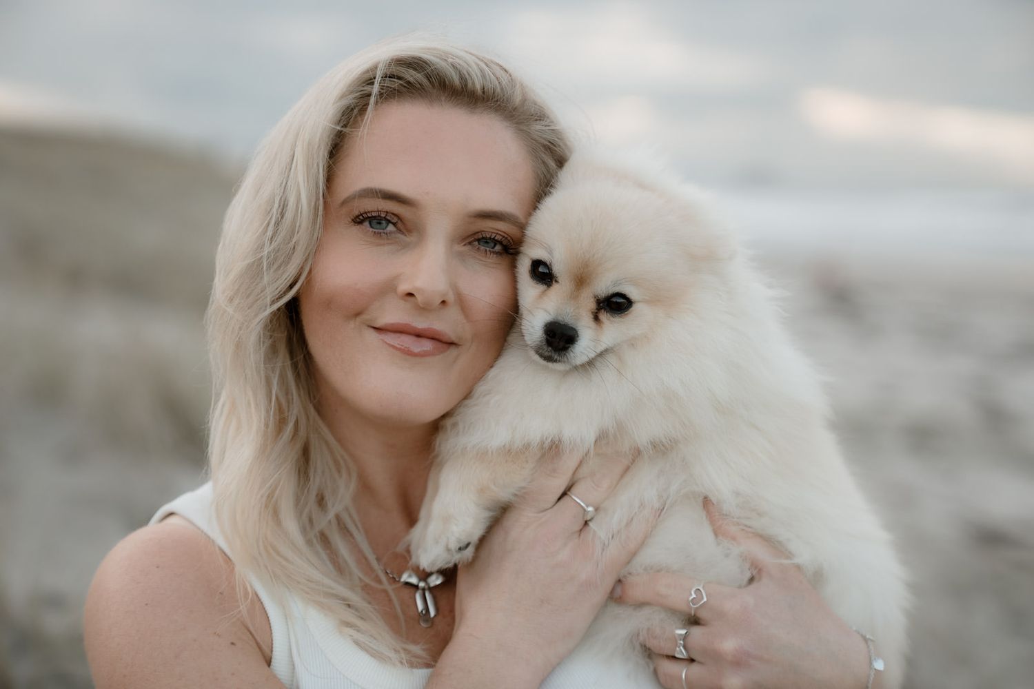 Smiling blonde woman holding a fluffy white Pomeranian puppy on a beach.