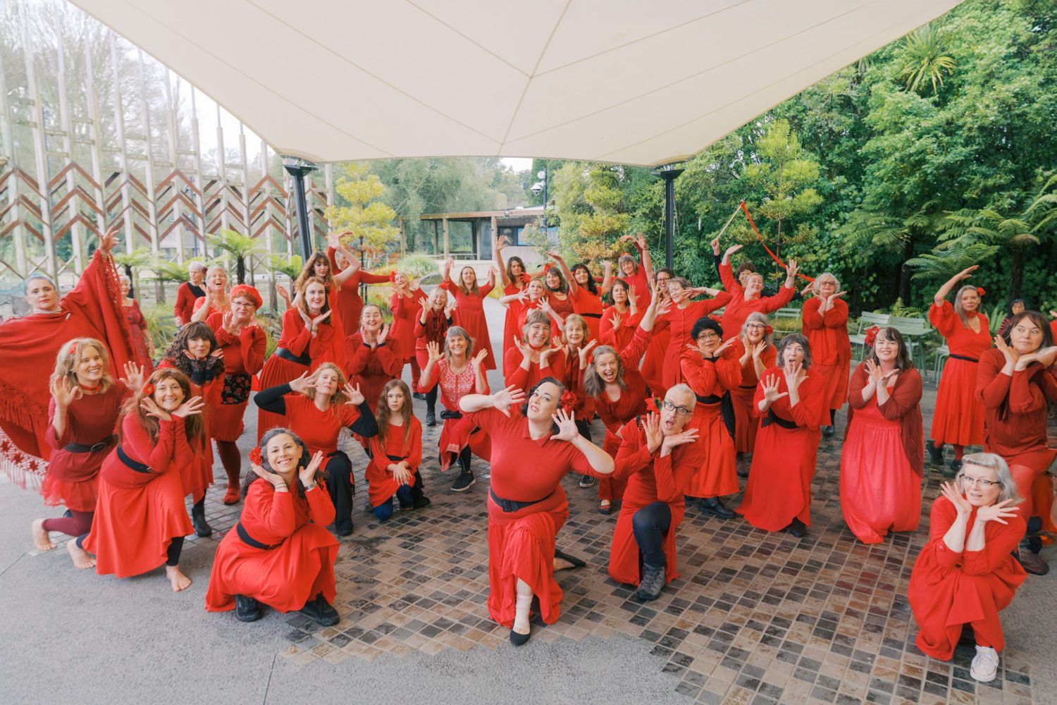 Large group of women in matching red dresses posing outdoors near a modern building surrounded by green trees.