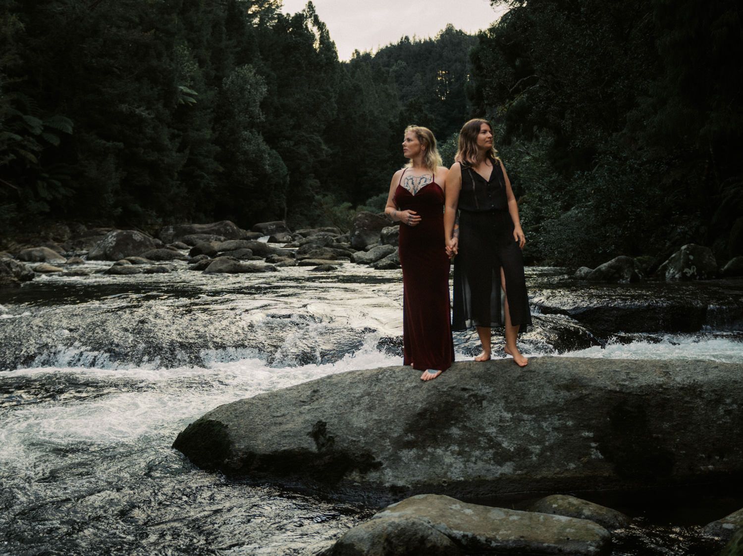 Two women in elegant gowns stand on river rocks amid flowing water and dense forest backdrop.