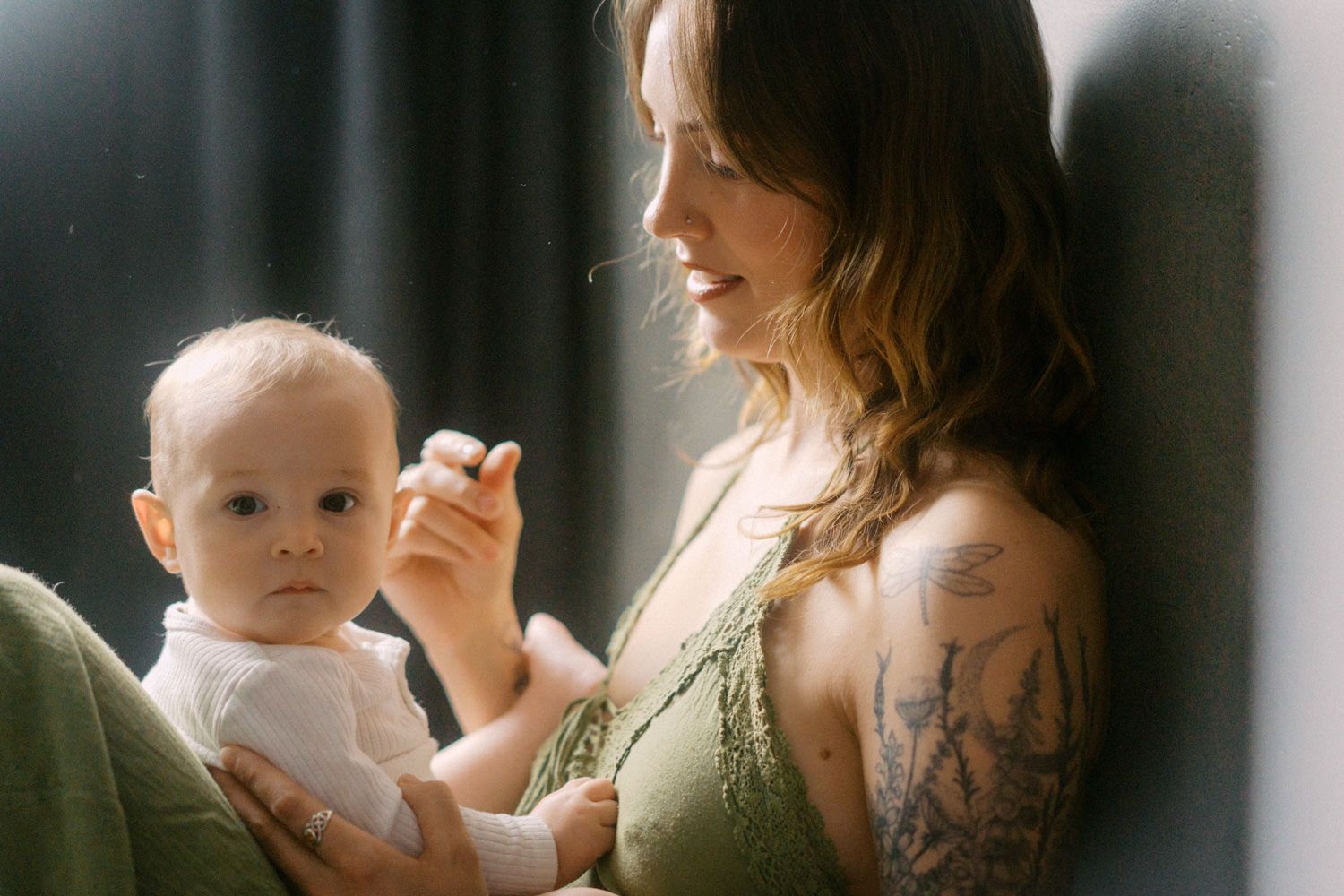 Tattooed mother tenderly holding her baby in soft natural light by a window.