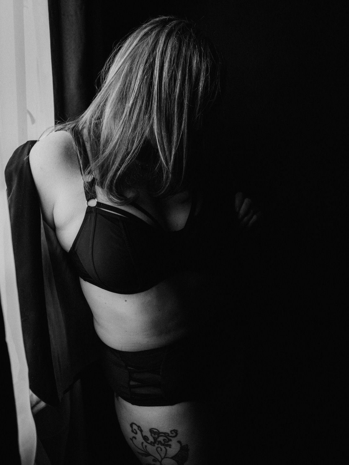 Black and white boudoir photo of a blonde woman in black lingerie with a thigh tattoo, posed by a window.